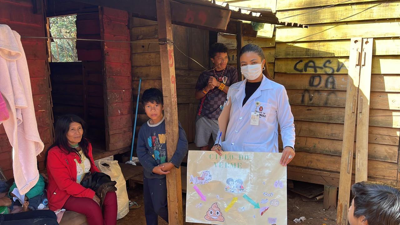 Na imagem, há quatro pessoas, sendo três indígenas (uma mulher, um adolescente e um menino) e a enfermeira Ednna, que segura um cartaz com orientações didáticas sobre parasitoses. Ela usa máscara de proteção e jaleco branco. Ao fundo, há uma casa de madeira